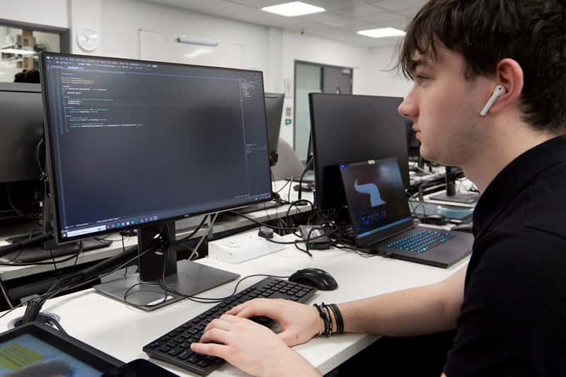 A photograph of a student with cropped black hair sat at a computer, writing lines of code on screen.