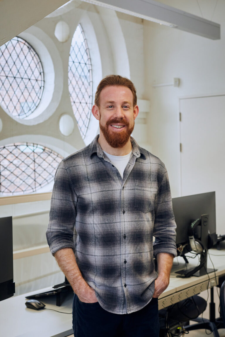 Staff profile image of Dan Kelby. Dan wears a plaid shirt over a white top, and is stood in front of the windows in the Media Lab