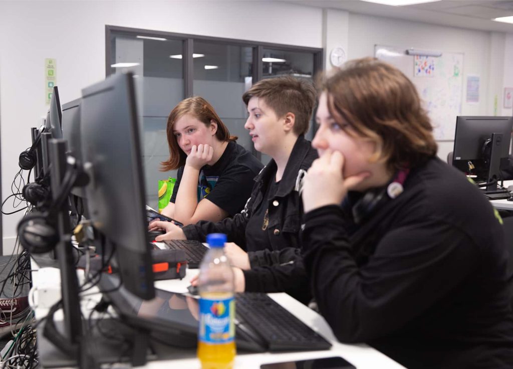 Three students sit in a row at computers. The photo is taken from the back, so we see the faces but not their screens. Two are interested in the same screen, while the third is concentrating on their own computer.