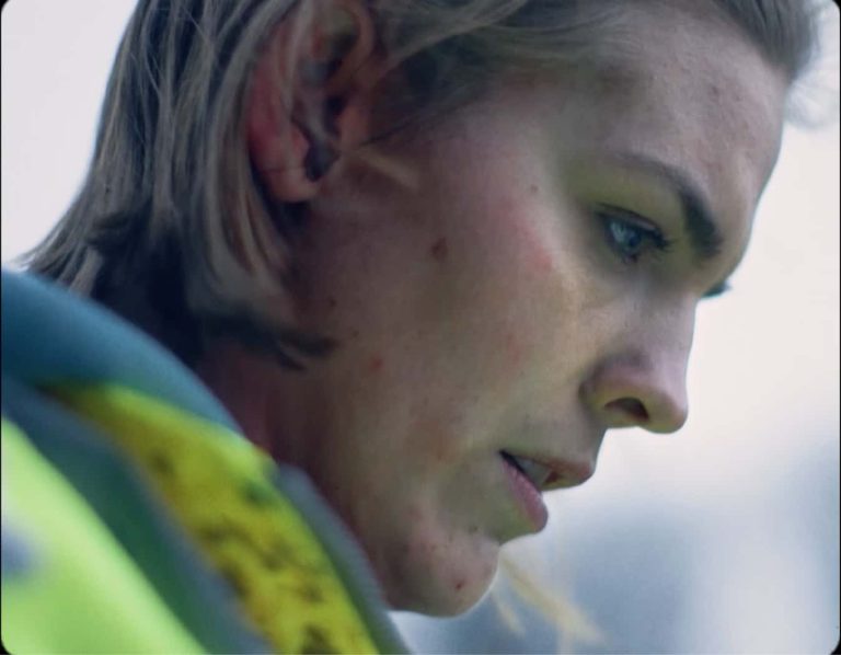 Still from film 'She Hears Crying' dir. George Irwin. Close up of a woman's profile, she has dark blonde hair and a bright yellow jacket.