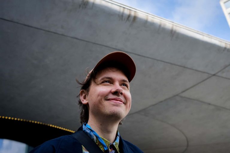 A portrait of Christian Petersen smiling and looking away from the camera. He is wearing a cap, blue shirt and jacket.