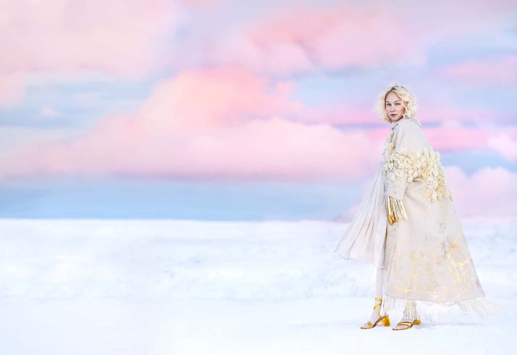 Image of a blonde haired model posing against a blue and pink sky stood on snowy ground.