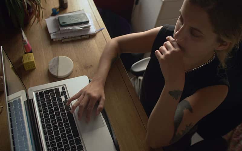 Still from film 'Is There Anybody Out There?' dir. Ella Glendining. Top down view of Ella Glendining, who sits at a desk with one hand on the trackpad of a laptop. She is wearing a black vest and has tattoos on one arm.