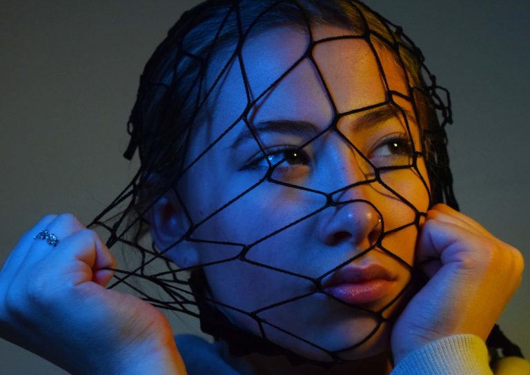 Close up of a young woman holding black fish netting over her face, with blue and orange lighting.