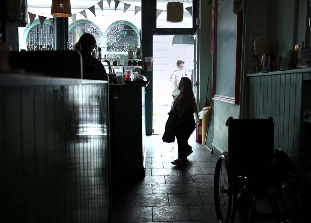 Still from film 'Is There Anybody Out There' dir. Ella Glendining. Silhouetted shot of a cafe, where one person is ordering at the till. There is a street outside and bunting on the window. A wheelchair is parked near the counter.