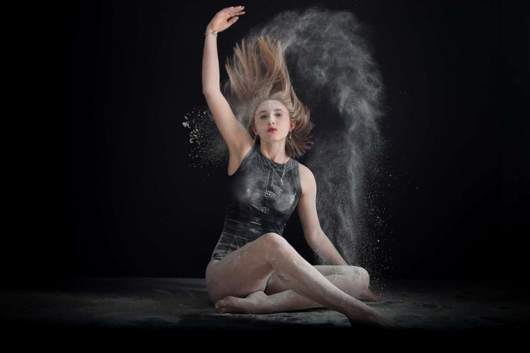 A young woman sitting against a black studio backdrop spraying white sand into the air.