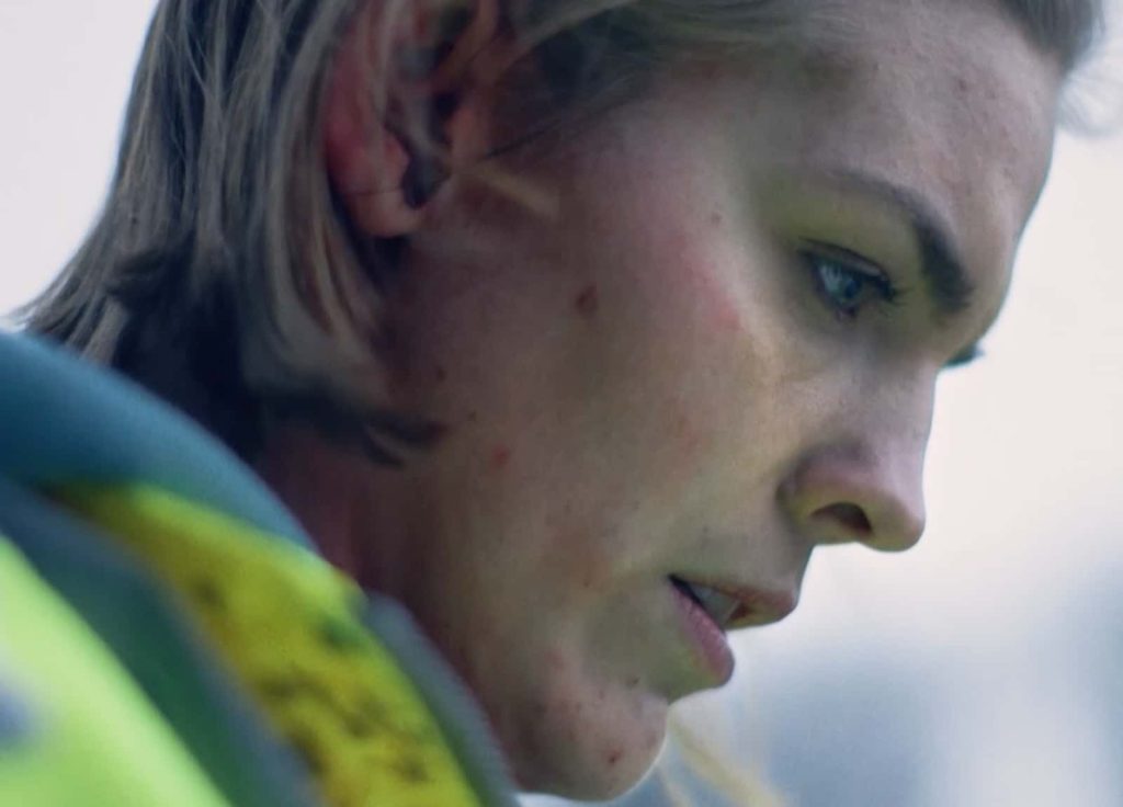 Still from film 'She Hears Crying' dir. George Irwin. Close up of a woman's profile, she has dark blonde hair and a bright yellow jacket.