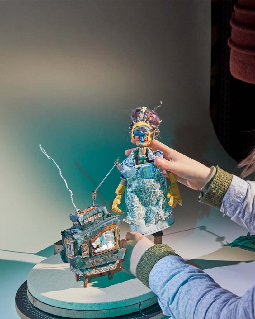 A close-up photograph of an animation student working in a studio environment. The work is a small stop motion character and tv.