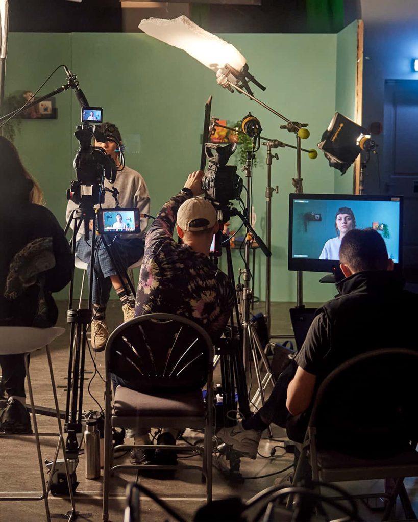 A photograph of a filmset with various lights, film cameras and monitors, that are pointed towards a woman sitting on a chair against a green background. The film crew includes 3 individuals that are all sat down behind the equipment.