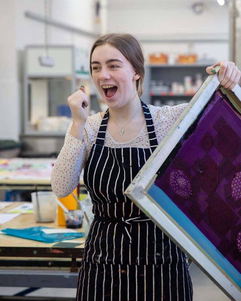 A female textile student smiling and raising her hand to gesture a moment of happiness as she uncovers her fabric from a print and dye workshop.