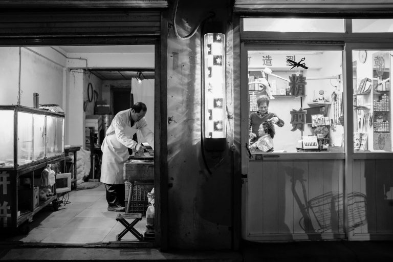 Black and white image looking into the window of a barber shop with a man cutting a customer's hair.