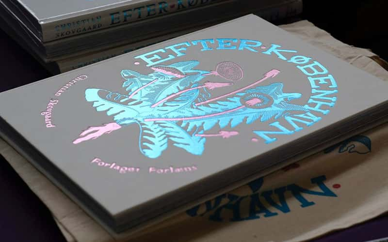 A hardback book laying flat. The book is named Efter København which is in large blue foil writing, along with a blue and pink foil plant illustration.