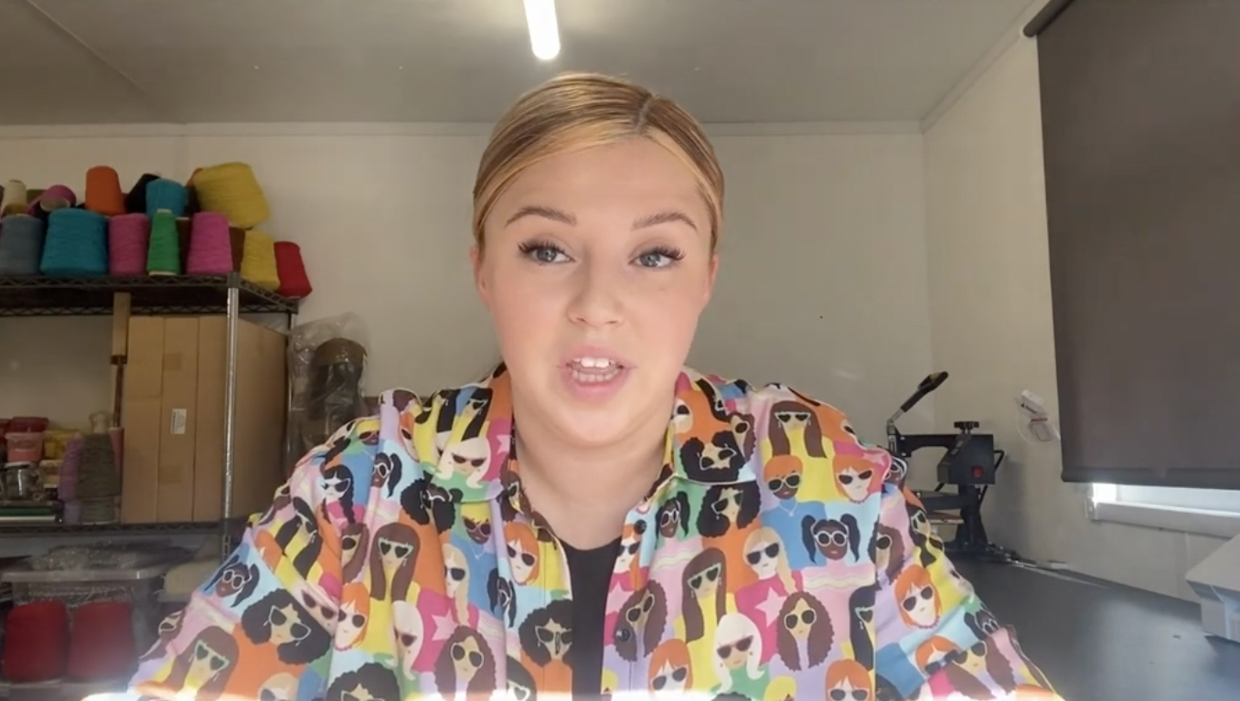 A woman with blonde hair tied back sits at a desk in a brightly lit room. She wears a colorful shirt with cartoon dog faces and looks toward the camera. Shelves with colorful yarn and supplies are visible in the background.