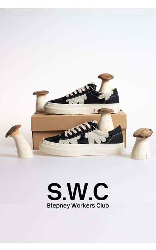 Stepney Workers club trainers photographed in a studio with a white background. One trainer sits on a cardboard box whilst the other sits underneath it with five woodland mushrooms surrounding the trainers.