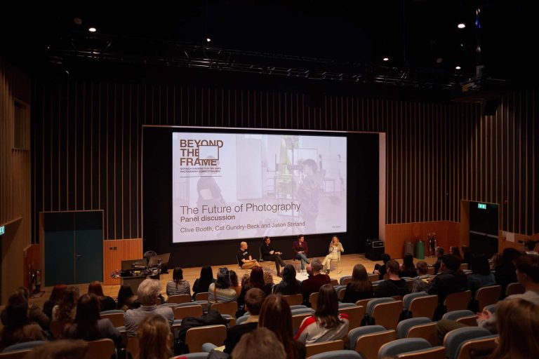 Photo from Beyond the Frame presentation. Shot from the back of a lecture theatre, the four judges are sitting on chairs in front of a large screen.