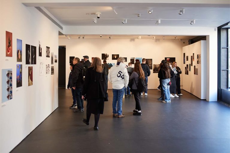 Wide shot of a gallery with lots of photographs hung on white walls. A crowd is looking at the pictures.