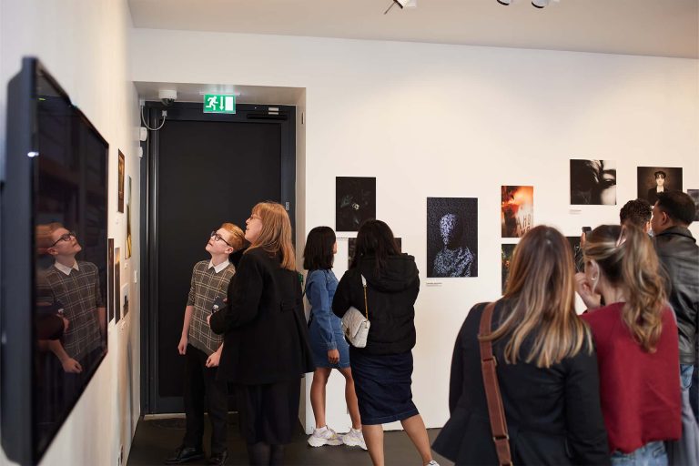 Wide shot of a gallery with lots of photographs hung on white walls. A crowd is looking at the pictures. A television is on the left wall.