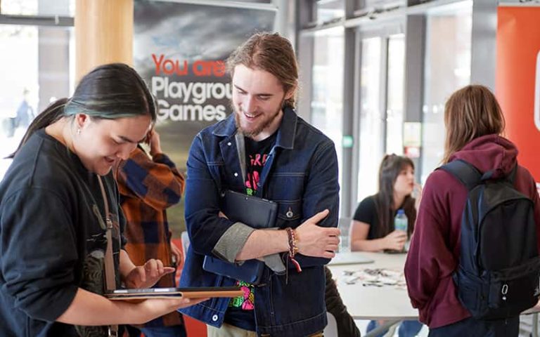 Two people with long hair are smiling at a tablet. People are gathered in the background around banners for Playground Games.