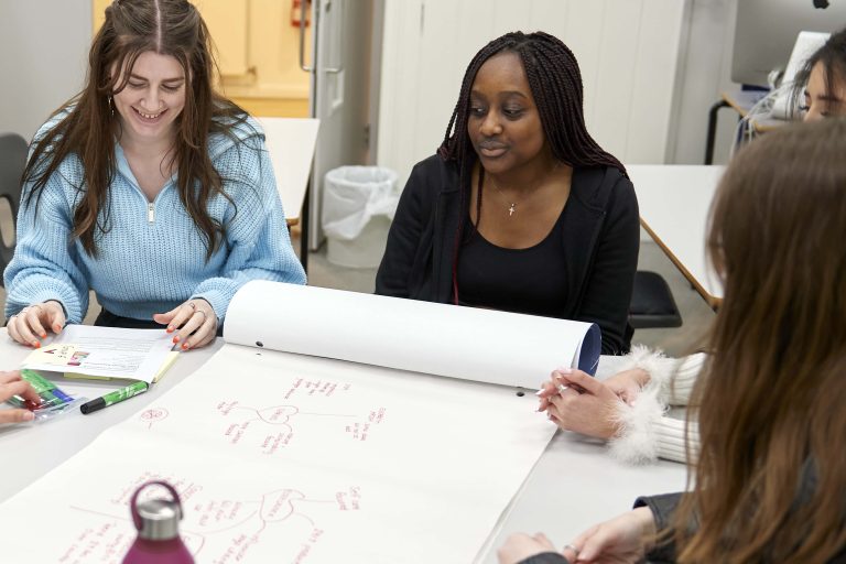 Two female students smiling and working together around a table. The table has a large piece of paper on it with a mind map drawing.