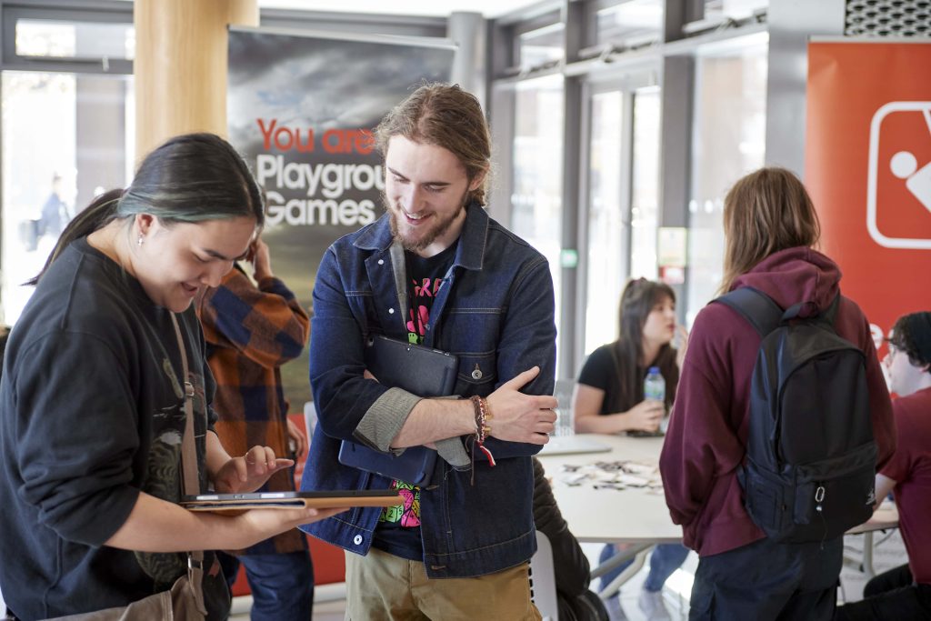 Two people with long hair are smiling at a tablet. People are gathered in the background around banners for Playground Games.