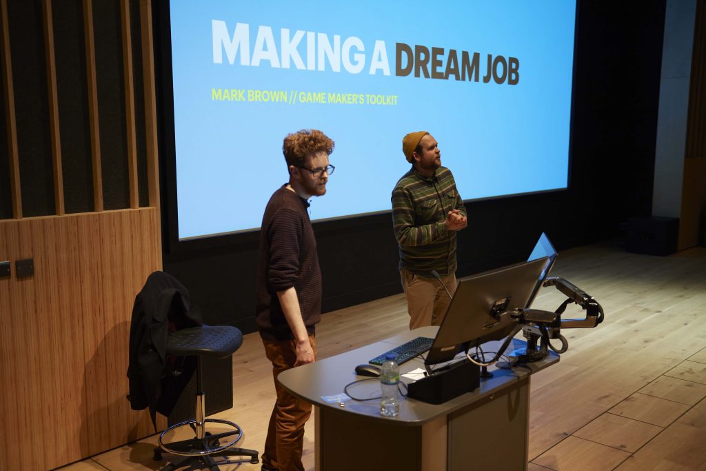 Guest speaker Mark Brown and Games Lecturer Jake Montanarini stand at the front of a lecture threatre. A big screen is behind them, with 'Making a Dream Job' written on a light blue background.