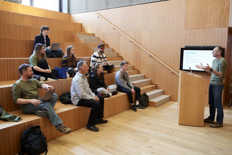 A speaker in a green shirt is presenting to a group of students. He is standing in front of a lectern, and the students are sitting on wooden, stepped seating.