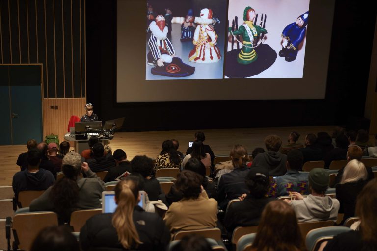 Visiting speaker Astrid Goldsmith is standing behind a lectern, presenting to a busy lecture theatre. There are pictures of small clay models on the big screen.