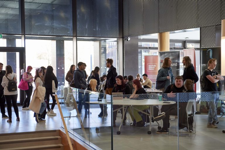 A busy foyer in a modern building with lots of wood and glass. People are busy chatting, some standing and some gathered around round tables.