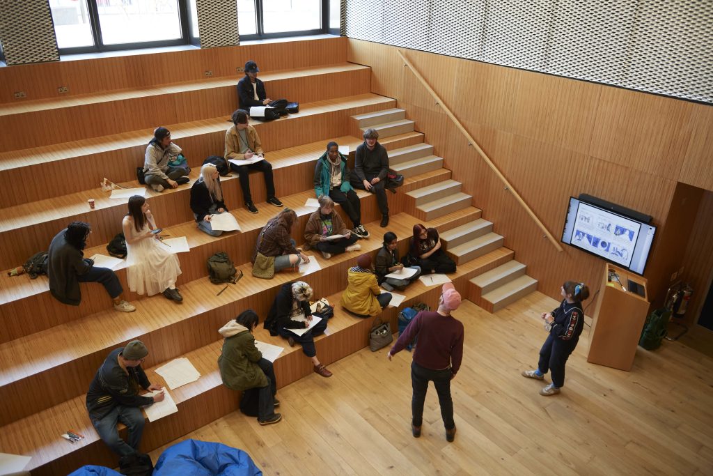 Top-down view of a storyboard workshop. Students are sat on a wooden stepped seating area, working on big pieces of paper. Two people stand at the front leading the session.