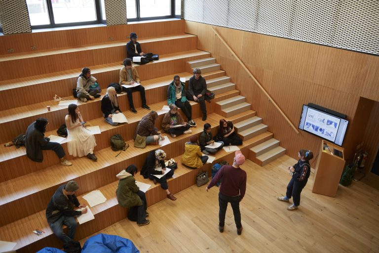 Top-down view of a storyboard workshop. Students are sat on a wooden stepped seating area, working on big pieces of paper. Two people stand at the front leading the session.