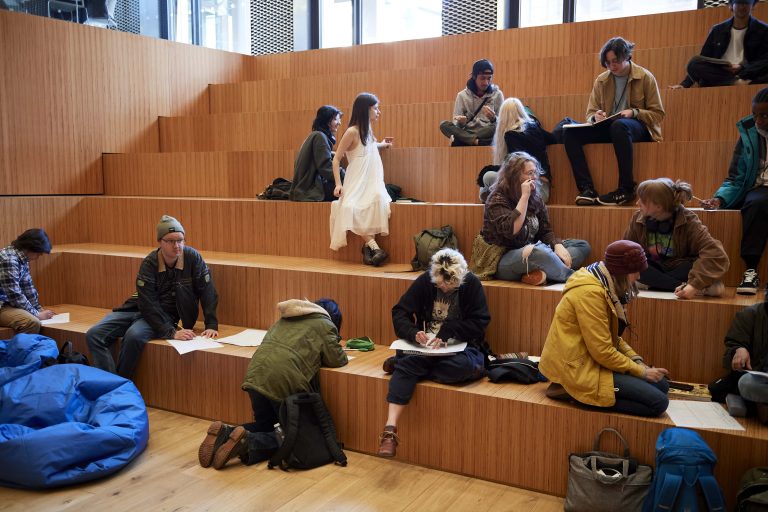 Students are sat on a wooden, stepped seating area, working on big pieces of paper.