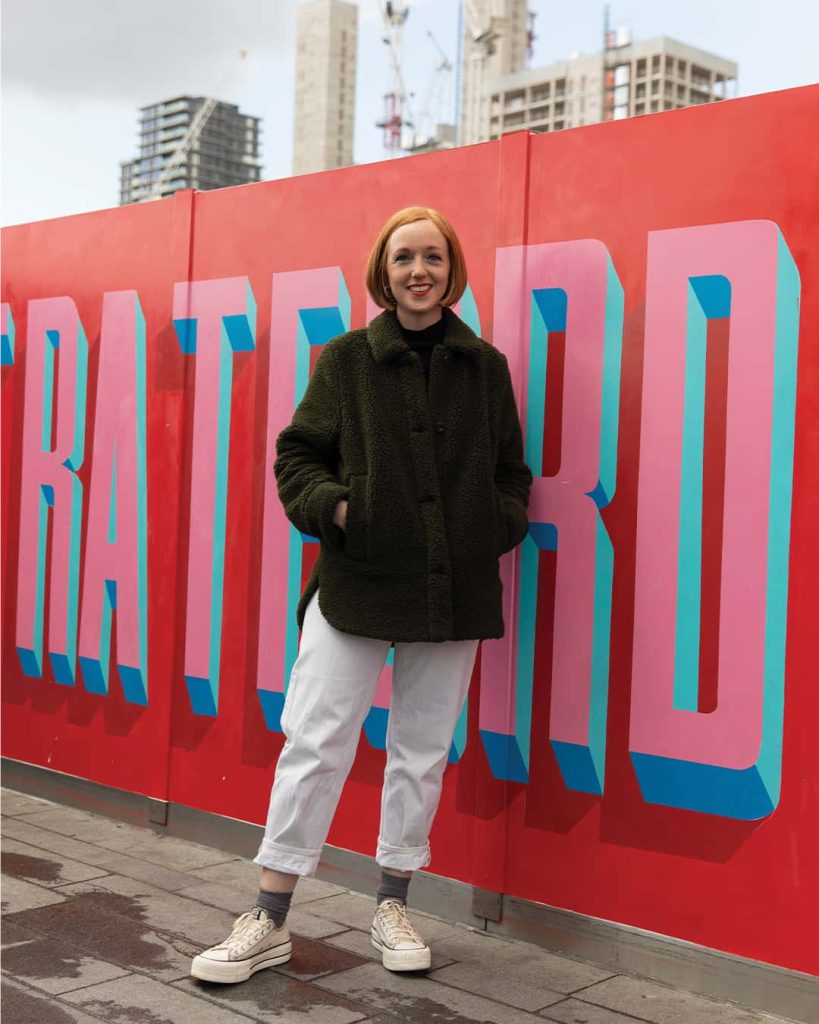 A full body portrait of Rachel Joy Price, a Graphic Design alumni. She is standing in front of a large outdoor canvas which she hand painted. The canvas has large letters on it spelling out the word 'Stratford' in red, pink and blue paint.