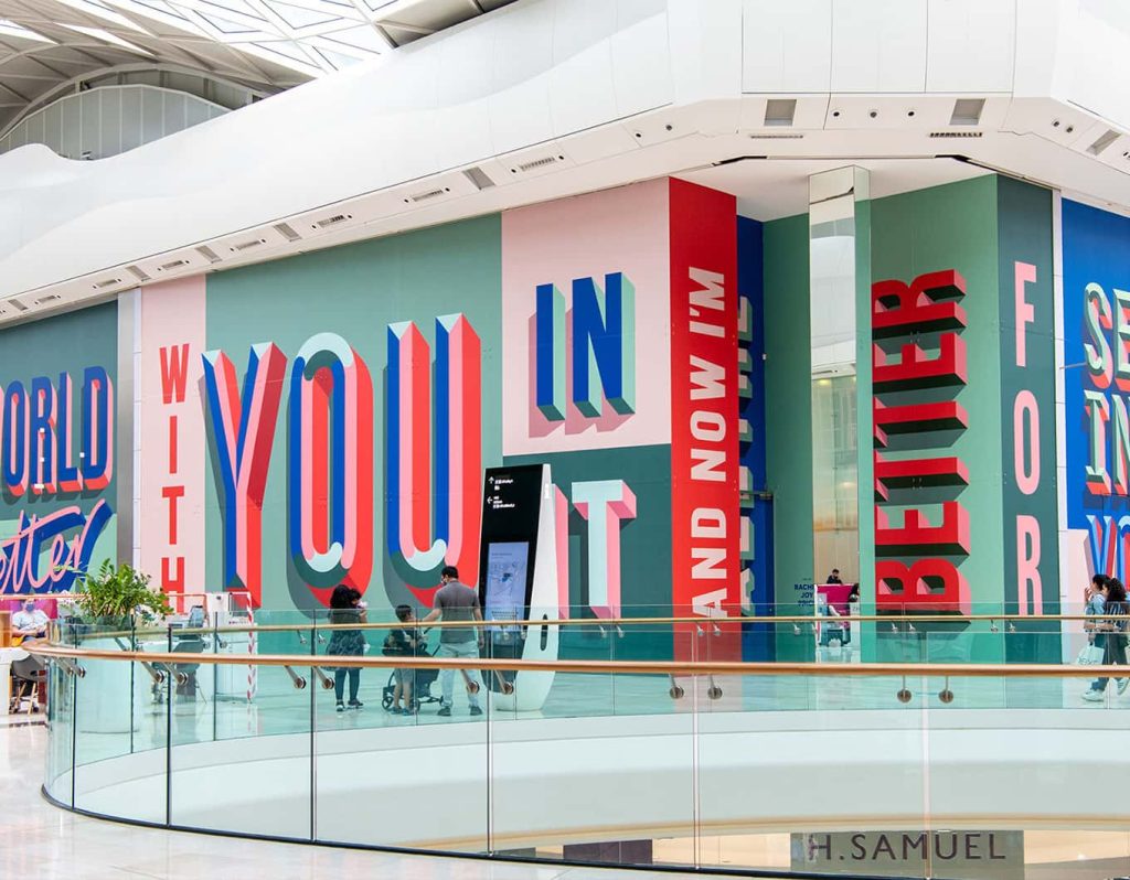 A photo of Rachel Joy Prices artwork in London Westfield shopping centre. The brightly coloured lettering has been printed onto empty shop units.