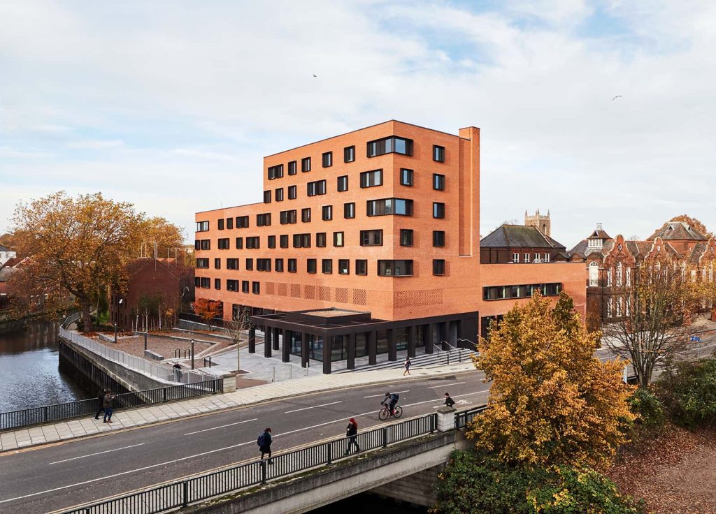 Duke Street Riverside accommodation at Norwich University of the Arts. A modern, orange brick building stands next to a river. A road bridge runs over the river, with people walking along the pavements.