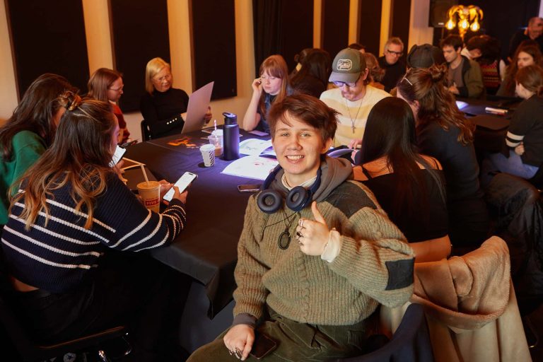 A smiling person in a grey jumper is thumbs up to the camera. Behind people are examining work around a round table.