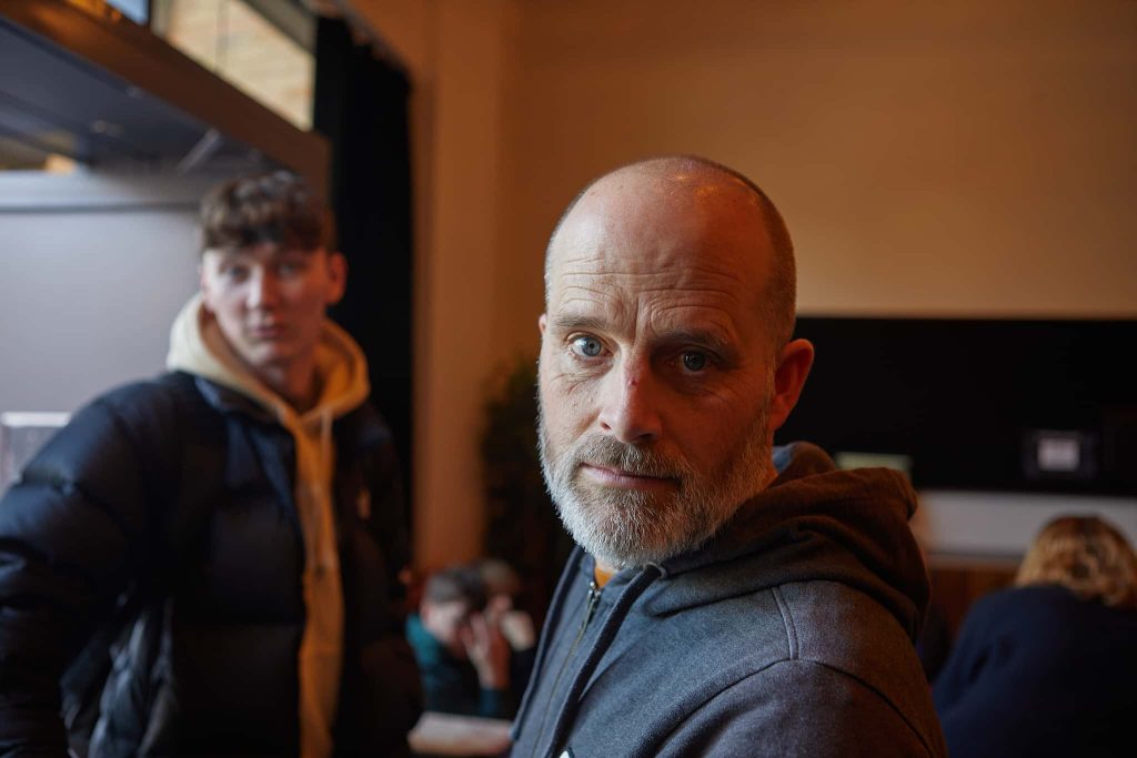 Head and shoulders shot of Photography course leader James Smith it looking right at the camera. To his right is a slightly out of focus person in the background with a navy coat and white hoody.