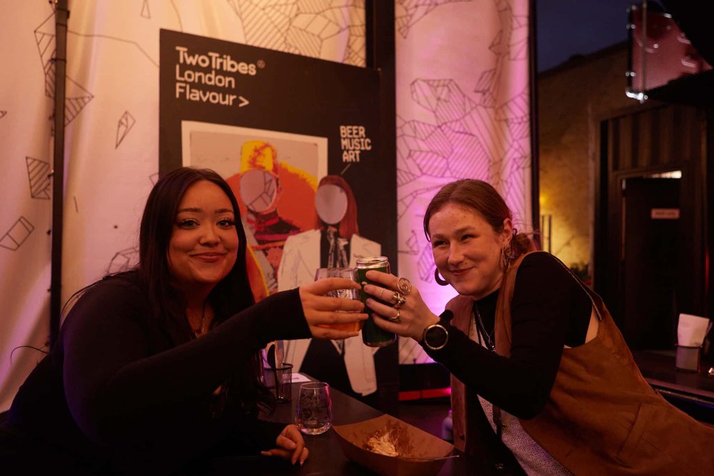 Two people are sat either side of a table smiling at the camera. They are 'cheers'ing their drinks over the table.