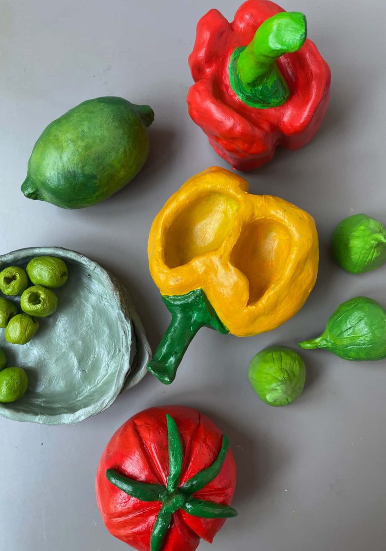 Bird's eye photograph of work by student Yasmin Shah. A collection of painted clay sculptures of fruits and vegetables, including a yellow pepper, a bowl of olives and a lime.