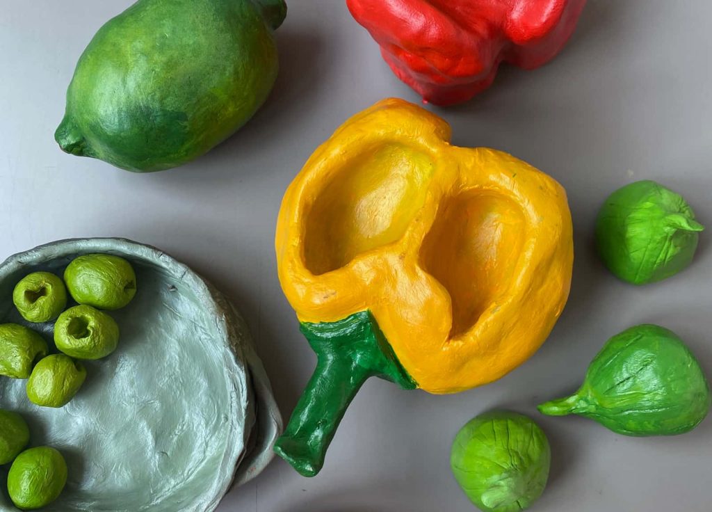 Bird's eye photograph of work by student Yasmin Shah. A collection of painted clay sculptures of fruits and vegetables, including a yellow pepper, a bowl of olives and a lime.