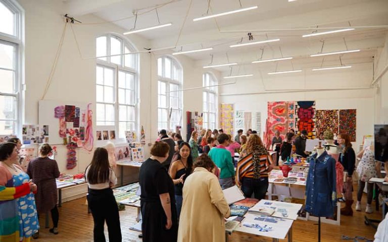 A photo of the BA (Hons) Textile Design Industry Awards. The photo is of students, judges and student work in a room where people are talking and looking at the student work that is laid out on tables and walls.