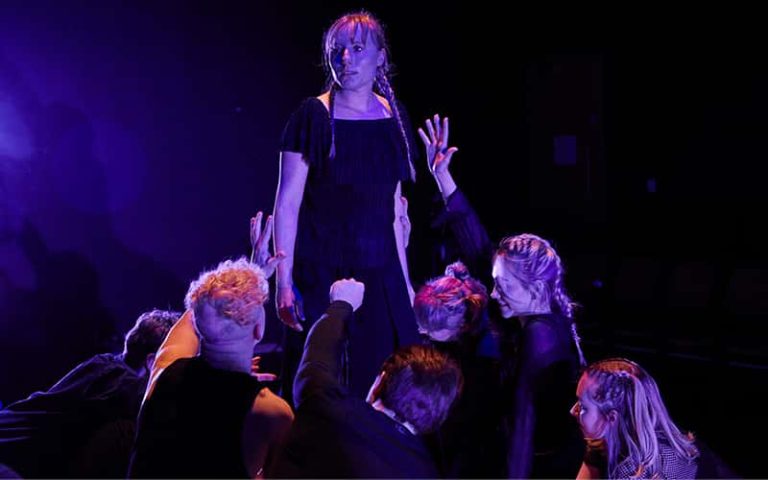 A woman stands raised above a group of people extending their hands towards her.