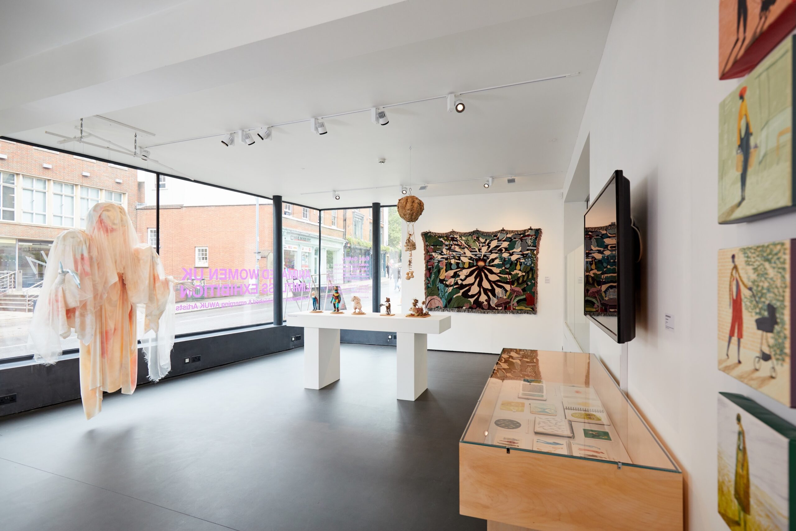 A bright contemporary art gallery with large windows, featuring sculptures on white pedestals, colorful wall hangings, paintings, a display case with documents, and a large textile figure near the window.