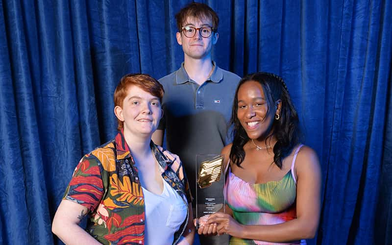 Three people are standing in front of a blue curtain, holding a glass award with a gold emblem on it.