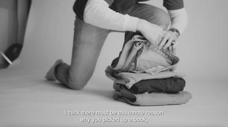 A black and white photograph of someone kneeing down to a stack of denim jeans.