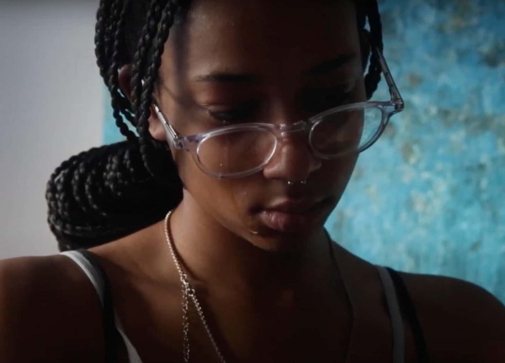 Still from film Under the Blanket (dir. Tanicha Toro-Oloto). Close shot of a person with long hair and glasses looks towards the floor, their face partly in silhouette.