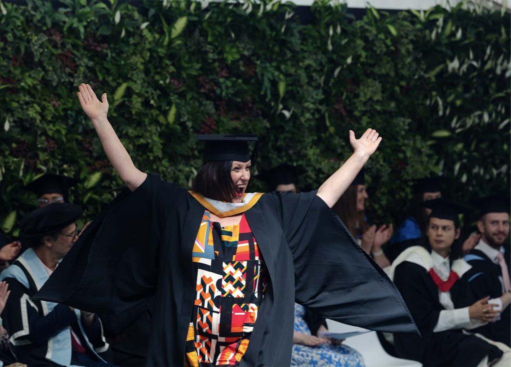 A photo of a student on stage at Graduation 2023 with their arms up in the air, celebrating as they walk across the stage.