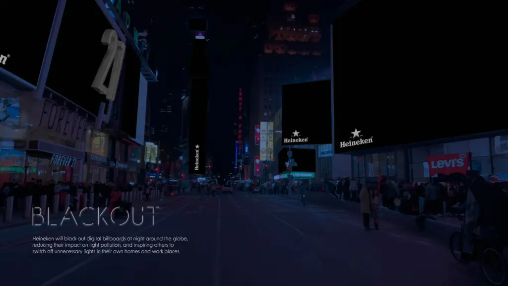 A dark city street at night with most digital billboards turned off, except a few displaying the Heineken logo. People stand along the sidewalks. Text at the bottom explains the "BLACKOUT" campaign to reduce light pollution.