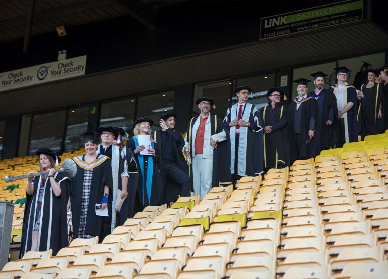 A photo of Norwich University of the Arts staff members, all dressed in graduation gowns as they wait on the stadium stairs for the procession to begin.