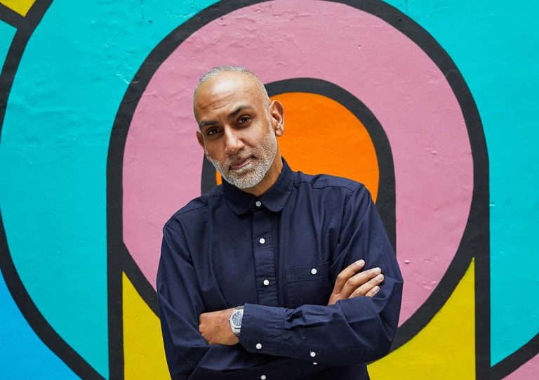 Gush Mundae wearing a navy shirt with crossed arms standing against a graphic mural.