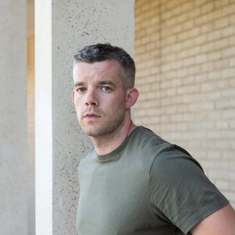 Russell Tovey wearing a green T Shirt standing in front of a column and brick wall.